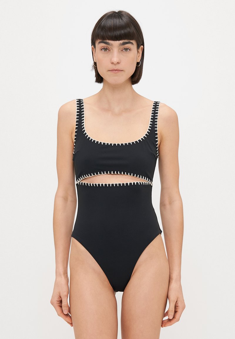 Čierne jednodielne plavky s výstrihom do lodičky, výrezmi a kontrastným prešitím pozdĺž okrajov a ramienok. Hladký povrch.