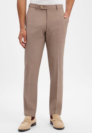 Mann trägt braune, maßgeschneiderte Hose und beige Loafers, steht vor schlichtem Hintergrund mit einer Hand in der Tasche.