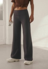 Platas kājas sporta bikses tumši pelēkā audumā ar gludu tekstūru, apvienotas ar brūnu saitē topu un baltām sporta apaviem. Ūdens pudele rokā.