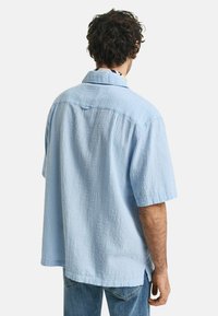 Camicia a maniche corte con bottoni, tessuto testurizzato azzurro chiaro, vestibilità rilassata, dettaglio sul retro, spacchi laterali, indossata con jeans azzurri chiari.