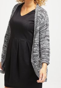Cardigan aperto in maglia bianco e nera. Maniche lunghe con tessuto testurizzato e vestibilità ampia. Abbinato a un abito nero con scollatura a V.
