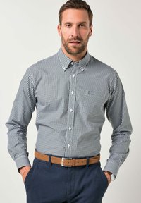 Camisa de botonera con un patrón de cuadros en azul marino y blanco, de mangas largas, cuello abotonado y un pequeño logo en el pecho. Metida dentro de pantalones oscuros.