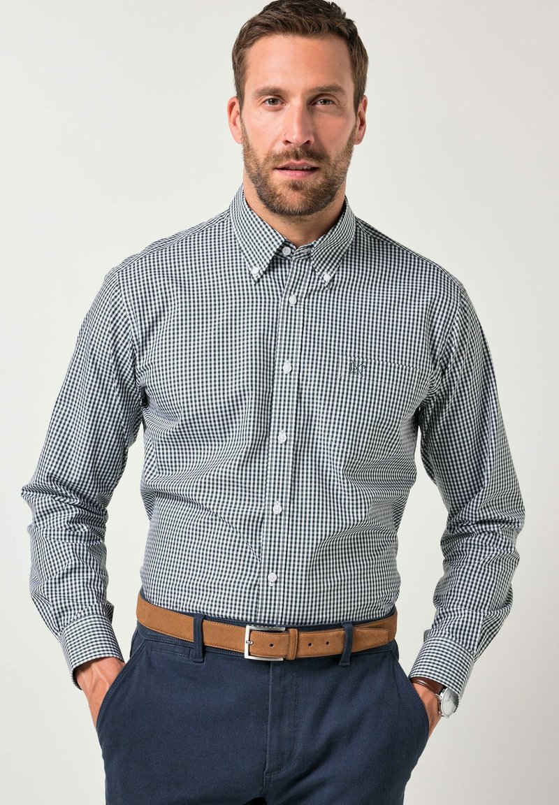 Camisa de botonera con un patrón de cuadros en azul marino y blanco, de mangas largas, cuello abotonado y un pequeño logo en el pecho. Metida dentro de pantalones oscuros.
