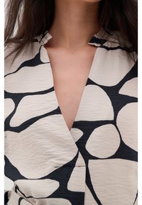 Femme portant un haut beige et noir à motif abstrait avec un décolleté en V profond et un col volanté, exposant une partie du cou et de la poitrine.