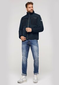 Marinejakke med høj krave, to brystlommer og lynlås foran; båret med lyseblå jeans og sportshvide sneakers.