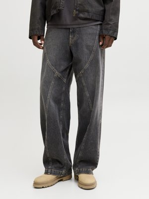 Osoba ubrana w luźne, ciemnoszare dżinsy z widocznymi przeszyciami, beżowe, solidne buty i czarną kurtkę na jednolitym tle.