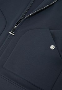 Indumento blu navy con una cerniera argento e una grande tasca con chiusura a bottone a pressione argento.