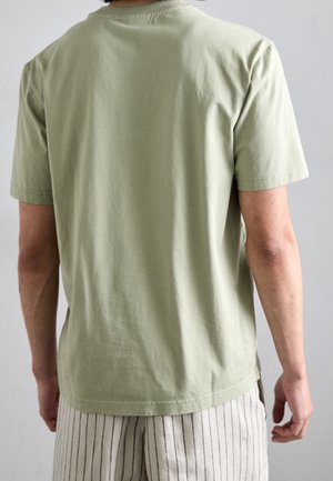 Jasnozielona bawełniana koszulka z krótkimi rękawami, z okrągłym dekoltem i gładką fakturą. Noszona z spodenkami w paski, widok od tyłu.