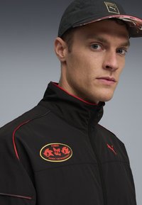 Puma-jakke i sort med røde detaljer, med lynlås foran og krave. Viser et mærke med Sonic the Hedgehog-design.