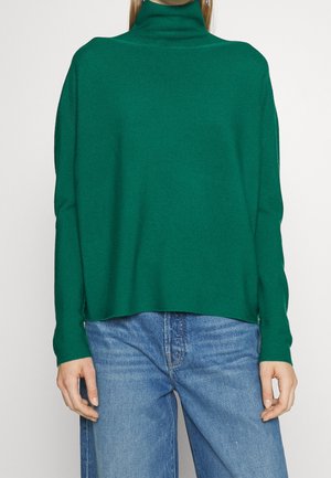Femme portant un pull à col roulé vert émeraude à manches longues et un jean taille haute bleu clair, visible du cou jusqu'aux hanches.