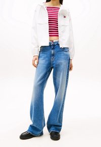 Witte capuchonjas over een rood-wit gestreept crop top, gecombineerd met lichtblauwe wijde jeans en zwarte schoenen.