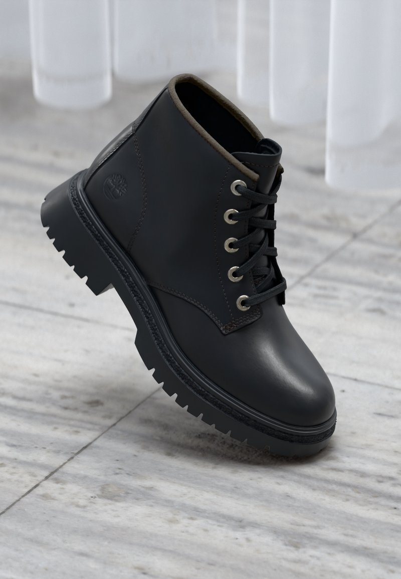 Bottine en cuir noir avec semelle à gros crampons et œillets en métal, photographiée sur un sol en marbre gris clair avec des rideaux blancs en arrière-plan.