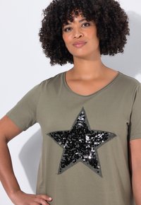 Camiseta verde oliva de manga corta con un gran diseño de estrella en lentejuelas negras y plateadas en la parte frontal. Textura de tela suave.