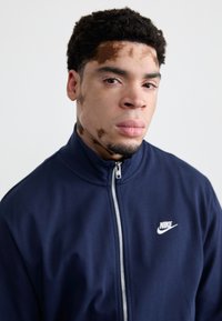 Casaco Nike azul-marinho com fecho e colarinho alto e logótipo branco no peito. Tecido suave com ligeiro brilho, design ajustado.