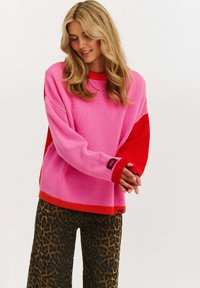 Jasnoróżowy i czerwony sweter o luźnym kroju, z prążkowanymi mankietami i dołem. Połączony z spodniami w panterkę. Widoczny design w blokach kolorystycznych.