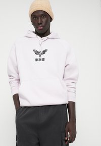 Αχνό ροζ φούτερ με μπροστινές τσέπες, γραφικό μαύρης πεταλούδας και ιαπωνικό κείμενο. Φορεμένο με σκούρα γκρι καμπαρντίνες και μπεζ σκουφί.