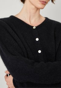 Melnā, pūkaina kardigāna ar V veida kakla izgriezumu un četriem apaļiem, baltajiem pogām priekšpusē. Tam ir garas, brīvi plūstošas piedurknes un maiga tekstūra.