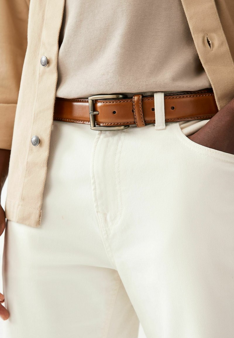 Cinturón de cuero marrón con hebilla metálica rectangular, usado sobre pantalones de tela clara y camisa beige, con una capa exterior beige y botones plateados.