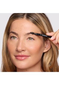 Femme aux cheveux châtain clair coiffant son sourcil avec une brosse à sourcils noire portant la mention « Tweezerman », sur un fond uni.