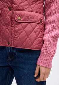 Ροζ μπουφάν Barbour με επένδυση σε σχήμα καπιτονέ και ορειχάλκινα κουμπιά, φορεμένο πάνω από ροζ πλεκτό πουλόβερ, συνδυασμένο με μπλε τζιν και χαλαρό χέρι στο πλάι.