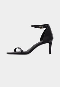 Sandale à talon haut noire pour femme avec fine bride à l’avant et bride cheville avec boucle dorée sur fond gris uni.