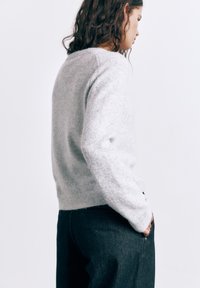 Žena s kučeravými vlasmi v svetlosivom svetri a tmavých nohaviciach, stojaca bokom s jednou rukou v vrecku na jednoduchom pozadí.