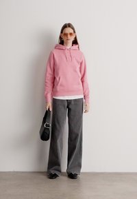 Sudadera rosa, camiseta blanca debajo, pantalones grises de pierna ancha, zapatos negros, sosteniendo un bolso negro. Accesorios: gafas de sol de color claro.