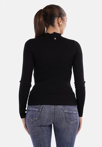 Maglione nero a coste con collo alto e maniche lunghe, caratterizzato da un piccolo dettaglio a forma di cuore dorato sul retro. Indossato con jeans blu.