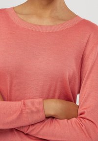 Růžový pletený svetr s ribbed kulatým výstřihem a manžetami na rukávech, který má jemnou texturu a uvolněný střih, zobrazený s překříženýma rukama.