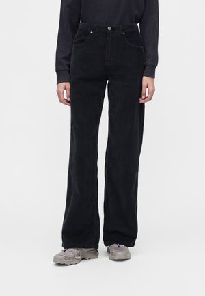 Personne portant un pantalon large en velours côtelé noir, un haut noir à manches longues et des baskets grises, debout sur un fond blanc.