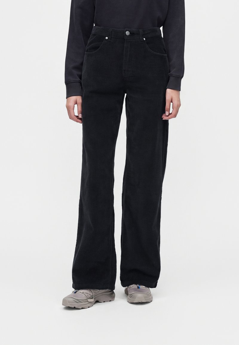 Personne portant un pantalon large en velours côtelé noir, un haut noir à manches longues et des baskets grises, debout sur un fond blanc.