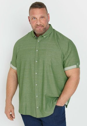 Mand med kort hår og skæg iført en grøn kortærmet skjorte med knapper og mørkeblå bukser, stående mod en hvid baggrund.