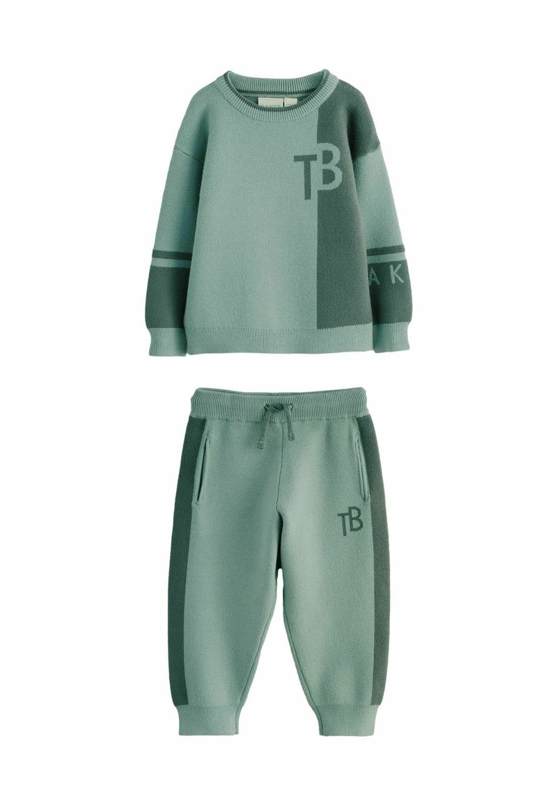 Minkštos žalios spalvos megzta dviejų dalių apranga, kurią sudaro megztinis su "TB" logotipu ir dryžuotomis detalėmis, derinama su atitinkamais joggeriais ir kišenėmis.