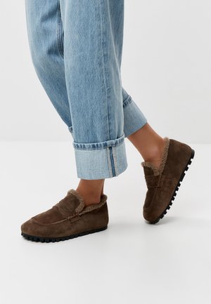 Noge u smeđim brušenim mokasinama obloženim toplim ovčjim krznom, uparene sa svijetloplavim trapericama s manžetama, na jednobojnoj pozadini.