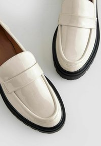 Mocassini in pelle verniciata crema con suola nera spessa, caratterizzati da una fascia frontale e design a punta arrotondata; finitura liscia con texture a contrasto.