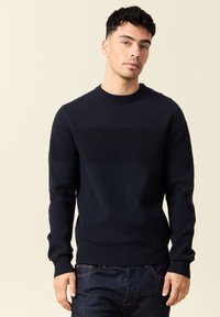 Maglione di colore navy in maglia con texture a coste orizzontali sul petto, colletto rotondo e polsini aderenti. Indossato con jeans scuri.