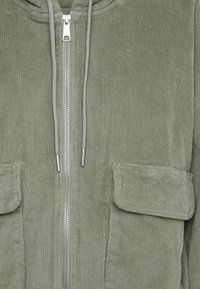 Kurtka sztruksowa w kolorze khaki z pełnym suwakiem, kapturem ze sznurkiem oraz dwoma przednimi kieszeniami. Charakteryzuje się teksturowanymi żeberkami i srebrnymi elementami wykończeniowymi.