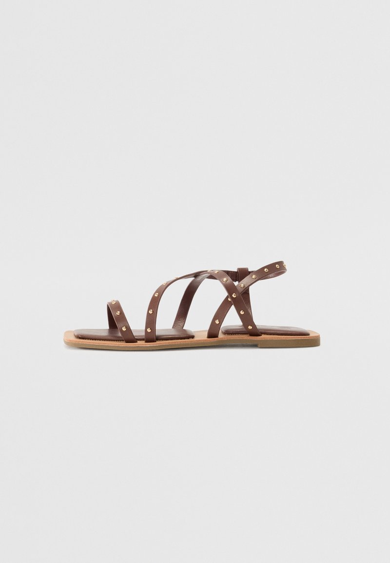 Brown leather sandals with multiple crisscross straps, accented by gold studs, flat sole, and minimalist design.