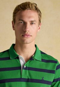 Zaļš un jūras zils svītrots polo krekls ar klasisku apkakli, kurai ir balts apakš-apkakle un neliels izšūts logo uz krūtīm.