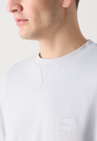 Sudadera gris claro con cuello redondo y un pequeño parche de logo texturizado. Presenta detalles de costura visibles y una textura de tela suave.
