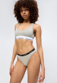 Γυναίκα με σγουρά μαλλιά που φορά γκρι αθλητικό σουτιέν Calvin Klein και ταιριαστό ριγωτό εσώρουχο, στέκεται μπροστά σε ουδέτερο φόντο.