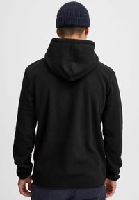 Schwarzer Fleece-Hoodie mit weicher Textur und einer verstellbaren Kapuze mit Kordelzug. Mit Rippbündchen an den Ärmeln und am Saum, getragen über einer dunklen Hose.