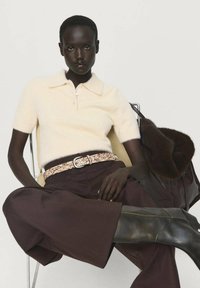 Modello seduto con una polo a maniche corte color crema, pantaloni ampi marrone scuro, cintura con stampa leopardata, stivali neri e una grande borsa di pelliccia scura.