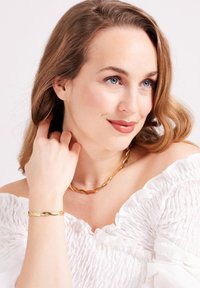 Goldene, gedrehte Halskette und Armband an einem Model, dazu ein strukturiertes, weißes Off-Shoulder-Oberteil. Die Haare in sanften Wellen gestylt, mit natürlichem Make-up.