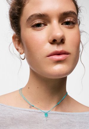 Joven con maquillaje ligero lleva pendientes de aro de plata y un collar fino de cordón turquesa con un pequeño colgante, sobre un fondo liso.