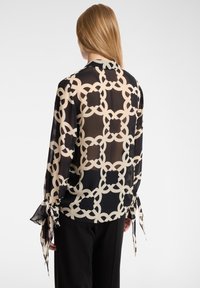 Durchsichtige schwarze Bluse mit einem cremefarbenen geometrischen Muster, ausgestattet mit langen Ärmeln und Schleifenakzenten an den Bündchen. Glatte Textur.