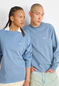 Two individuals wear blue long-sleeve shirts, one with a white collar. Both shirts feature a small "obey" logo on the chest.