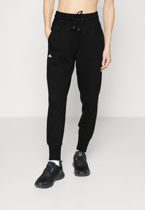 Personne portant un pantalon de jogging noir avec une taille à cordon et un logo blanc sur la cuisse gauche, assorti de baskets noires et de chaussettes blanches.