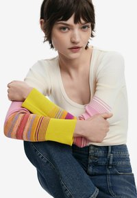Cremfarbener, gerippter Langarmoberteil mit bunt gestreiften Ärmeln in Pink, Gelb und Orange. Kombiniert mit dunkelblauen Jeans.