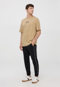 Beżowy t-shirt bawełniany z napisem "California", połączony z czarnymi dresami z białymi paskami po bokach i białymi trampkami z czarnymi akcentami.